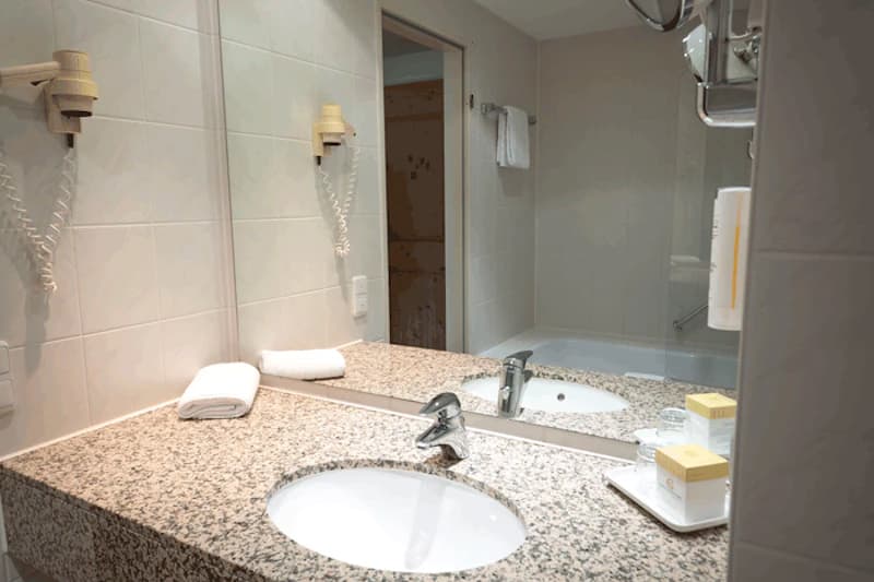 Hotelbadezimmer mit Waschbecken auf Granitablage, Spiegel, Wandfön, Handtücher und Badewanne mit Glasabtrennung.