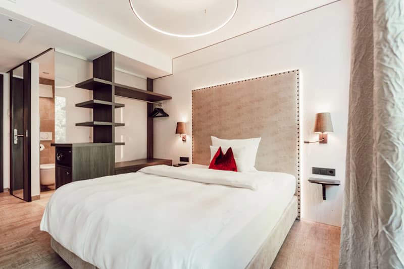 Doppelzimmer mit großem Bett, beleuchtetem Kopfteil, rotem Zierkissen, offener Garderobe und Blick ins Bad.