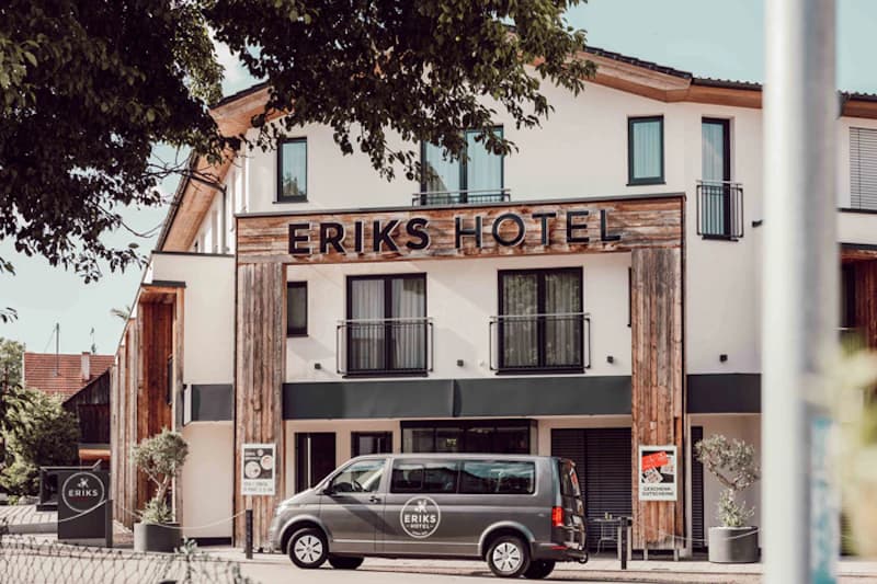 Fassade von Eriks Hotel mit Holzverkleidung, Schriftzug, Balkonfenstern und grauem Hotelbus vor dem Eingang.