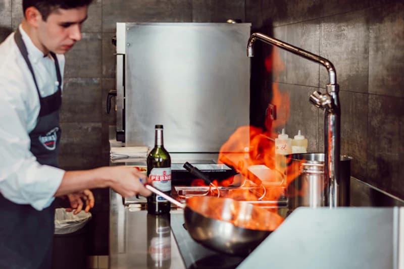 Koch in Schürze hält Pfanne mit hohen Flammen in einer Edelstahlküche neben einer Peroni-Flasche.