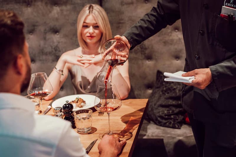 Kellner gießt Rotwein in Glas während Paar an Restauranttisch sitzt.