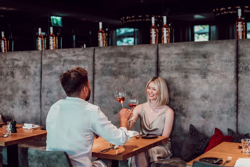 Paar stößt mit Weingläsern in einer Hotelbar-Sitznische mit grauem Polster an.