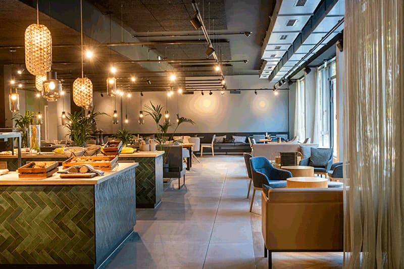Hotelrestaurant mit Buffetinseln, Hängeleuchten, Sitzgruppen, Pflanzen und großen Fenstern mit Vorhängen.