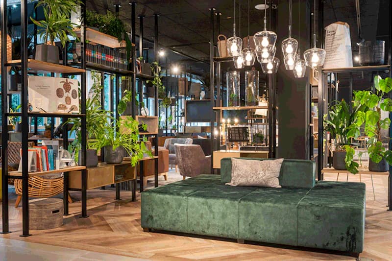 Offener Hotel-Loungebereich mit grünem Samtsofa, Regalen, Pflanzen und Hängeleuchten.