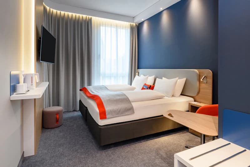 Hotelzimmer mit Doppelbett, blauer Akzentwand, Fernseher, Schreibtisch und großer Gardine.
