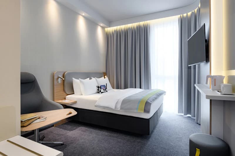 Modernes Hotelzimmer mit Doppelbett, Lesesessel, Wandfernseher, Teekanne auf Ablage und bodentiefer Vorhang.