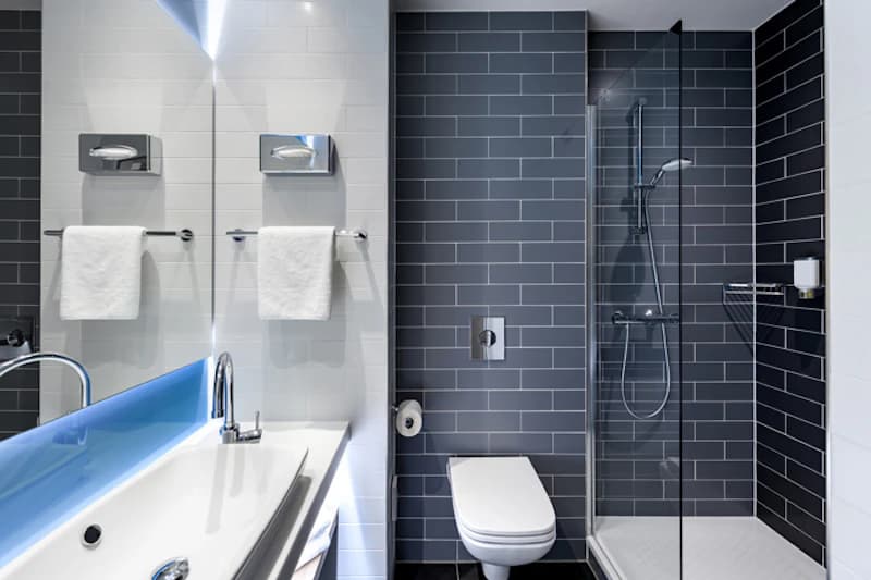 Hotelbadezimmer mit Waschbecken, beleuchtetem Spiegel, Handtüchern, Toilette und Glasdusche mit dunkelgrauen Wandfliesen.