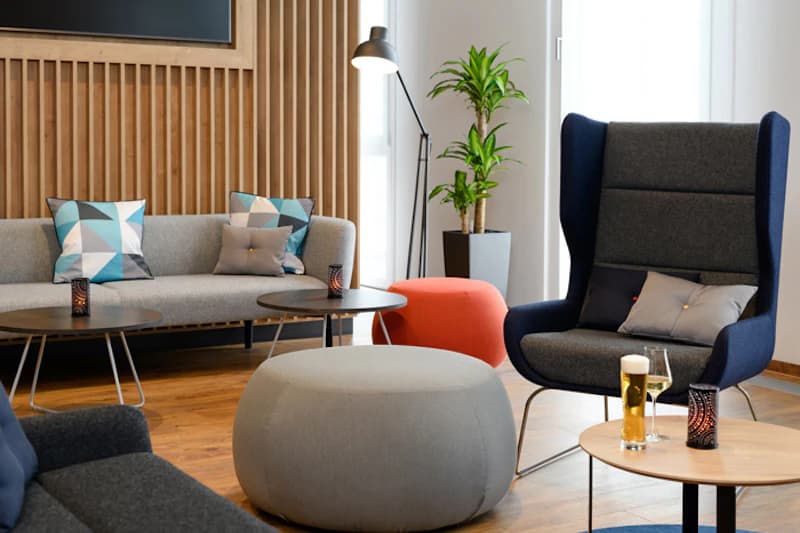 Moderne Hotellounge mit Sofa, Ohrensessel, runden Hockern, Beistelltischen, Stehlampe und Zimmerpflanze.