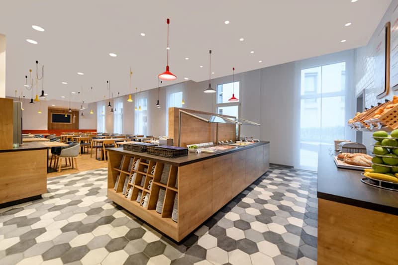 Frühstücksraum mit zentraler Buffetinsel, Regalen mit Tellern, Tischen, Stühlen und mehreren Hängeleuchten.
