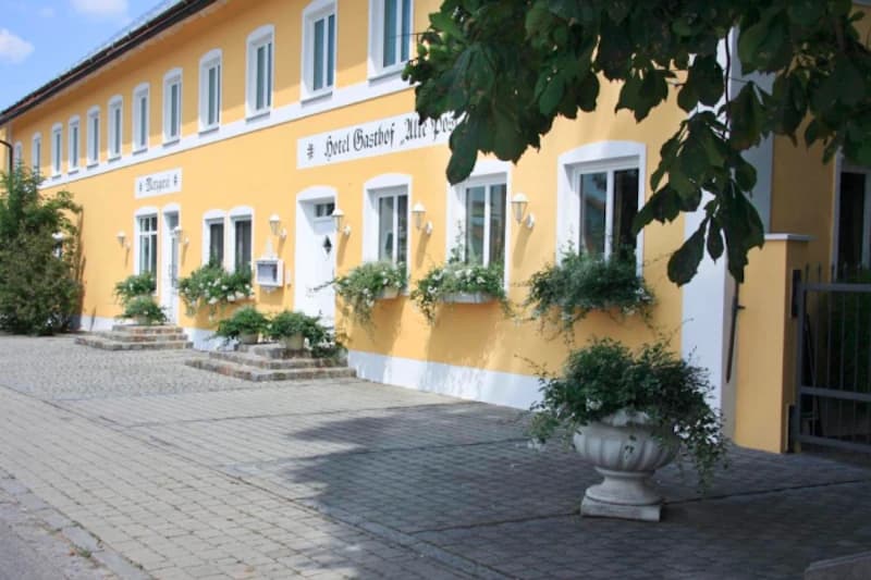 Gelbes zweigeschossiges Gasthofgebäude mit weißen Fensterrahmen, Blumenkästen an den Fenstern und gepflastertem Vorplatz.