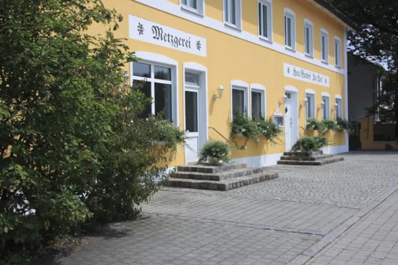 Gelbes zweigeschossiges Gebäude mit Eingangstufen, Fensterkästen mit Pflanzen und Schriftzug Metzgerei.