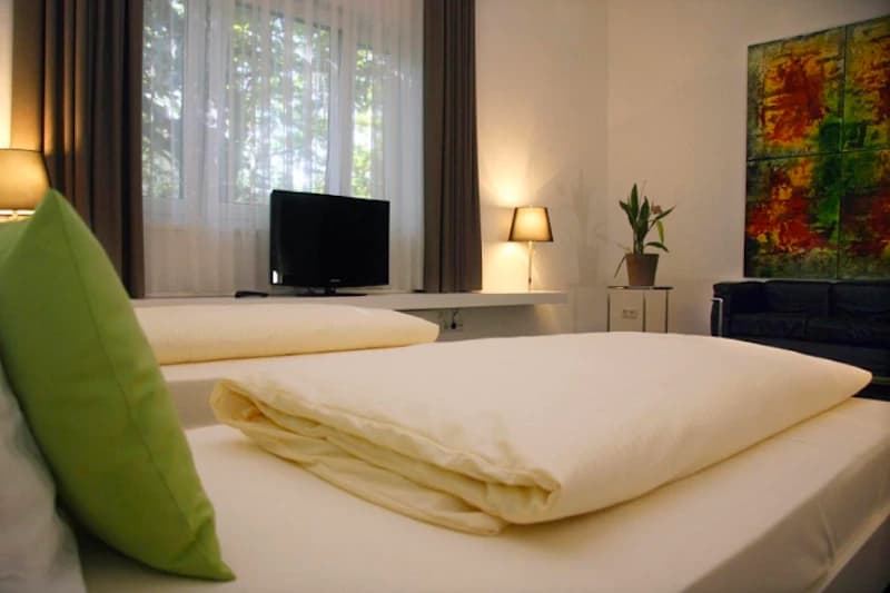 Zweibettzimmer mit weißen Bettdecken, grünem Kissen, Flachbildfernseher, Stehlampen, Sofa und abstraktem Bild.