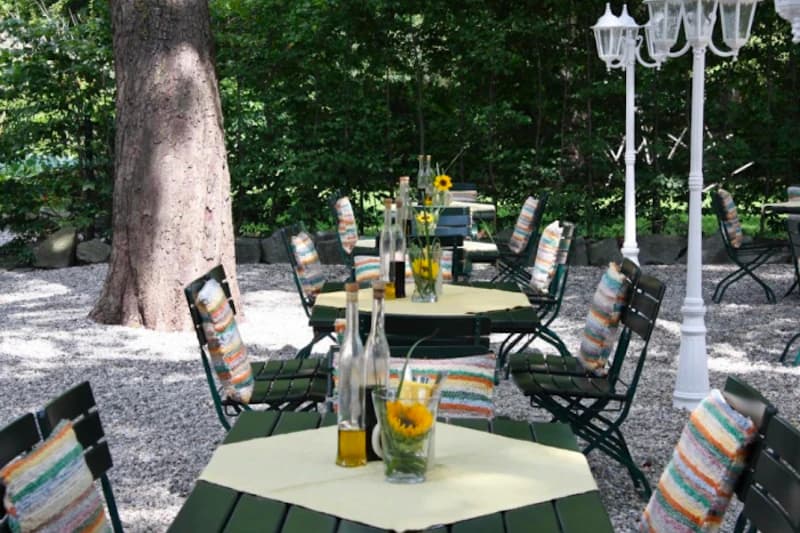 Grüner Außenbereich mit Tischen und Stühlen, gestreiften Sitzkissen, Sonnenblumenvasen auf Tischtüchern und Kiesboden unter Bäumen.