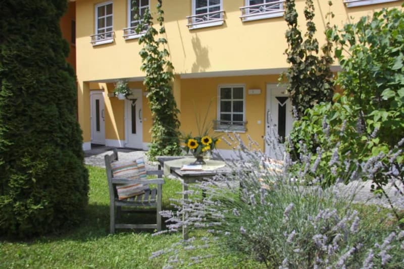 Gartensitzplatz mit Holztisch, Stühlen, Sonnenblumen vor gelbem Hotelgebäude.
