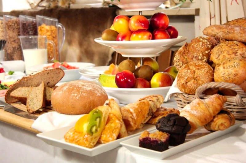 Frühstücksbuffet mit Brot, Körnerbrötchen, Croissants, Gebäck, Obst und Müslispender auf gedecktem Tisch.