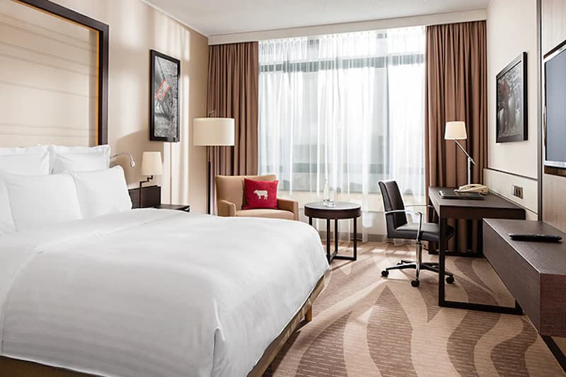 Hotelzimmer mit großem Bett, Sessel mit rotem Kissen, Schreibtisch, Flachbildschirm und großem Fenster mit Vorhängen.