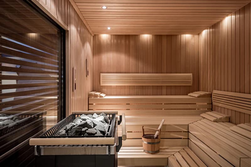 Finnische Sauna mit gestuften Holzbänken, Saunaofen mit Steinen und Holzeimer neben Glasfenster.