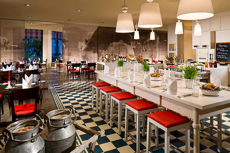 Helles Hotelrestaurant mit langer weißer Tafel, roten Hockern, Schachbrettboden und Buffetstationen.