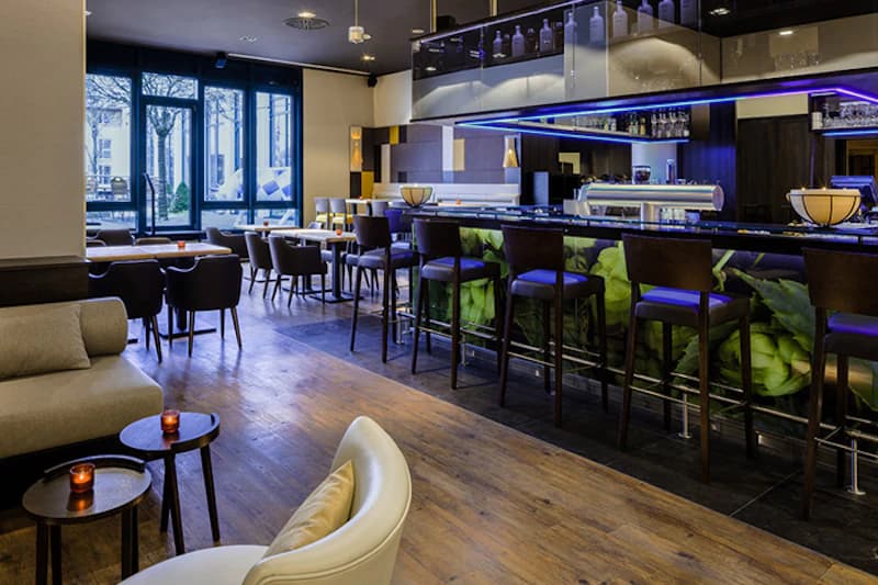 Hotelbar mit Theke und hohen Barhockern, Sitzgruppen und großen Fenstern zur Außenseite.