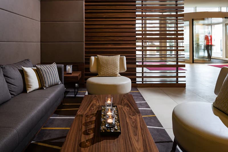 Hotelhalle mit Sofa, zwei Sesseln, Holztisch mit Kerzen und Lamellenraumteiler, Drehtür am Eingang im Hintergrund.