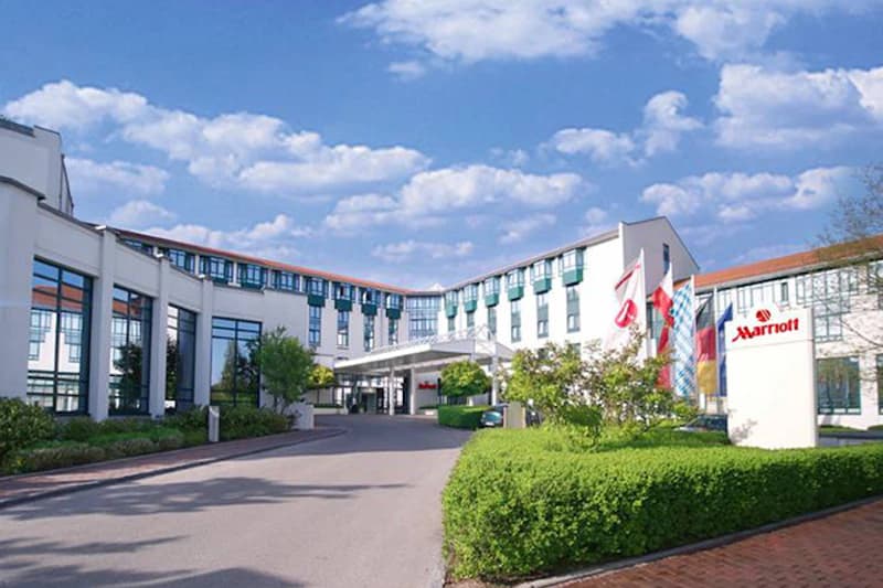 Eingangsbereich des Marriott Munich Airport Hotels mit Zufahrt, Vordach, Fahnen und Schildern neben gepflegten Hecken.