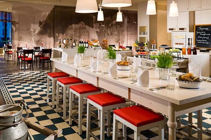 Restaurant mit langer weißer Tafel, roten Hockern, Buffet, kariertem Boden und hängenden Lampen.