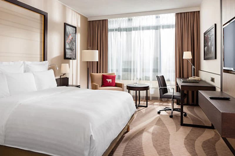 Helles Hotelzimmer mit großem Doppelbett, Sessel, Schreibtisch, Flachbildfernseher und Fenster mit Vorhängen.