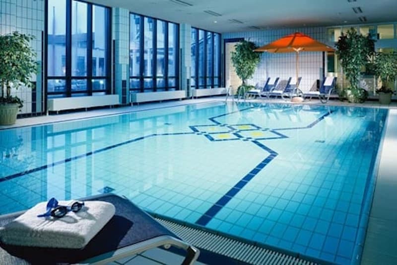 Innen-Schwimmbad mit blauem Becken, Liegen, orangem Sonnenschirm und Handtuch mit Schwimmbrille.