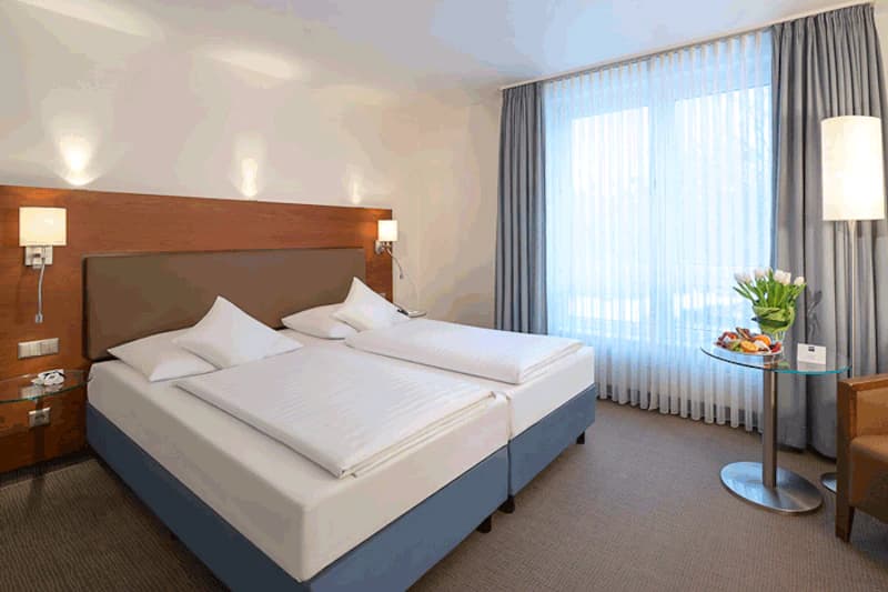 Hotelzimmer mit zwei Einzelbetten nebeneinander, Holzkopfteil, Nachttischlampen und großem Fenster.