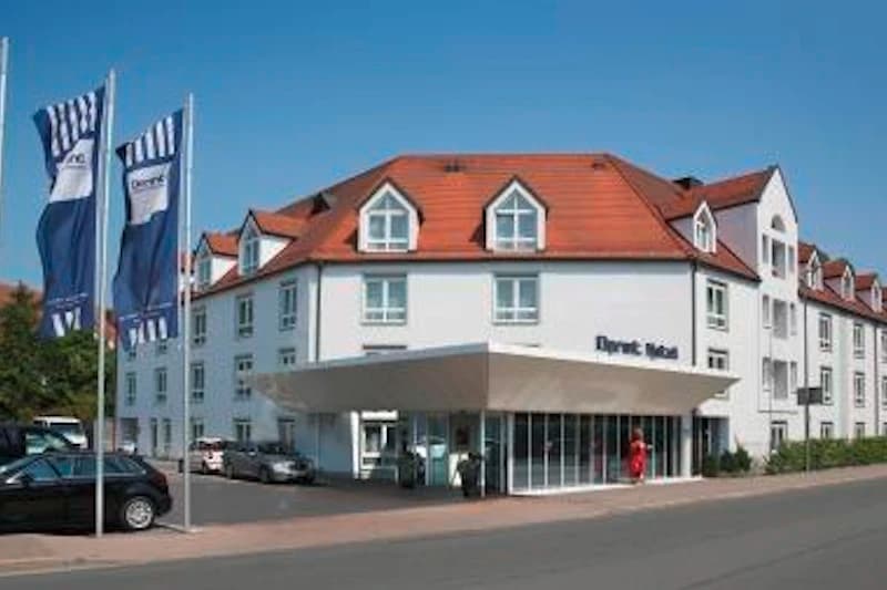 Dreistöckiges weißes Hotel mit rotem Walmdach, Eingangsüberdachung, drei Flaggen und mehreren geparkten Autos.