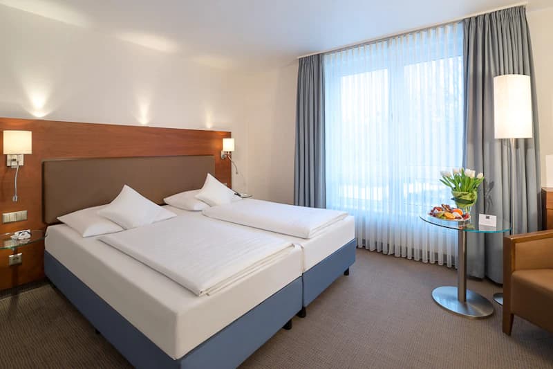Hotelzimmer mit zwei Einzelbetten nebeneinander, Holzkopfteil, Nachttischlampen und Rundtisch mit Blumen und Obst.