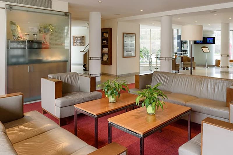 Hotelfoyer mit grauen Sofas, zwei Holztischen mit grünen Topfpflanzen und weißen Säulen.