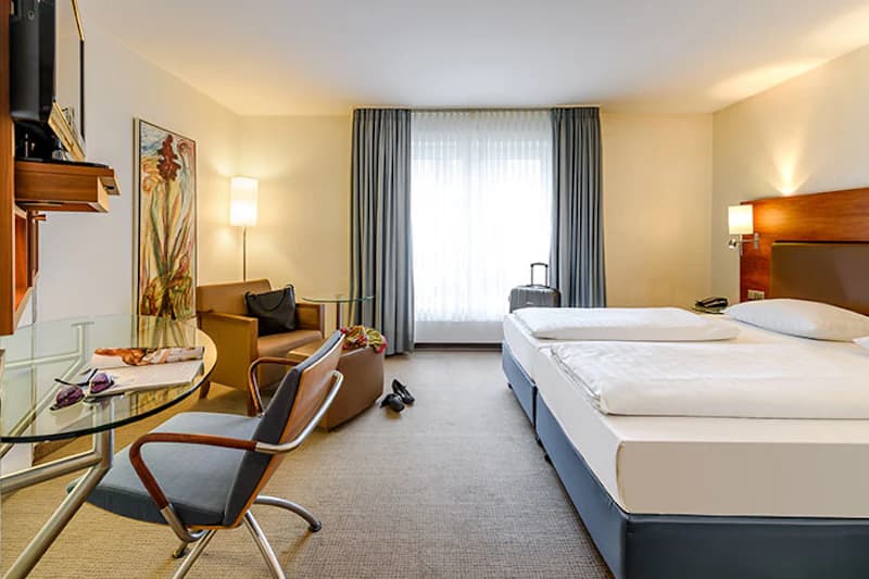 Hotelzimmer mit zwei Betten, Glasschreibtisch, Sessel, Stehlampe und Koffer am Fenster.