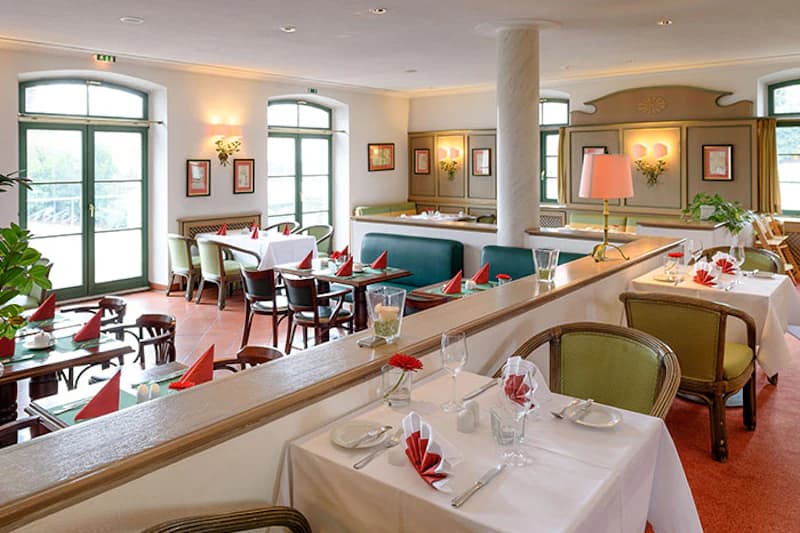 Helles Hotelrestaurant mit gedeckten Tischen, weißen Tischdecken und roten Servietten, Stühlen, großen Fenstern und einer Säule.