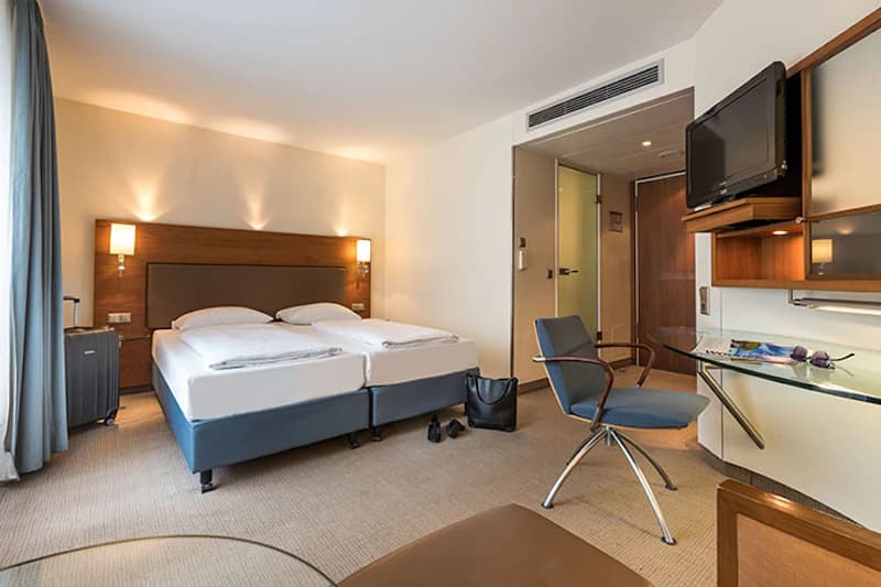 Hotelzimmer mit zwei Einzelbetten, Wandlampen, Schreibtisch, Fernseher, Stuhl, Koffer und Tasche.