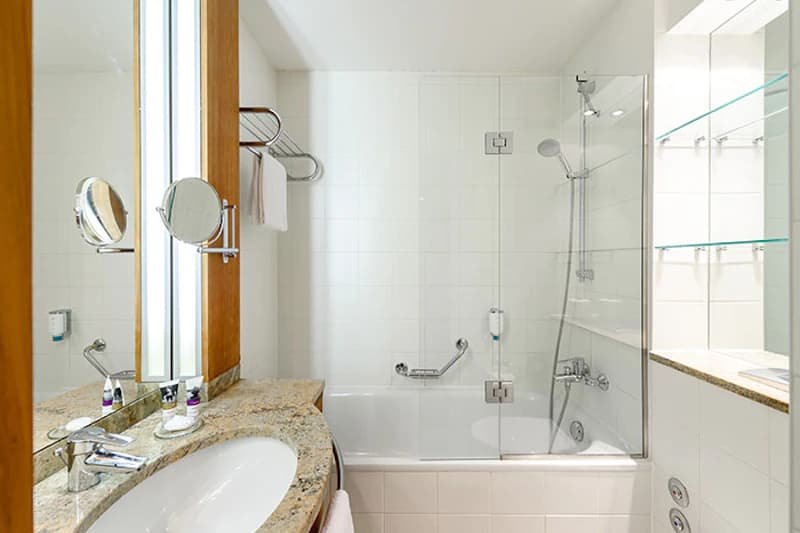 Hotelbadezimmer mit Waschbecken, Marmorplatte, beleuchtetem Spiegel, Badewanne mit Glasduschabtrennung und Handbrause.