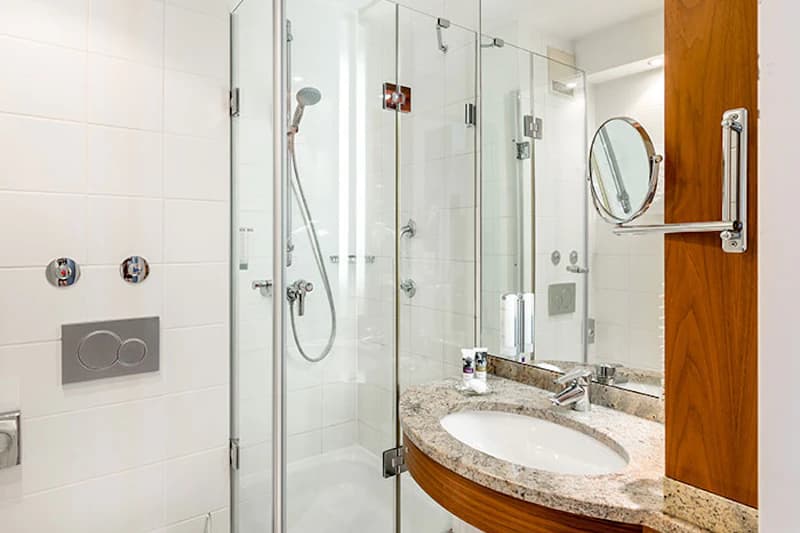 Kompaktes Hotelbad mit Glasdusche, Handbrause, Waschbecken mit Granitplatte, Spiegel und Holzschrank.