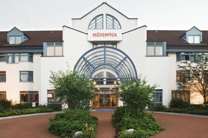 Hoteleingang des Mövenpick Hotels am Flughafen mit geschwungener Glasüberdachung, Zufahrt und Vorgarten.