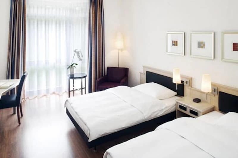 Zweibettzimmer mit zwei Einzelbetten, Nachttischlampen, Sessel, Schreibtisch und großem Fenster mit Vorhängen.