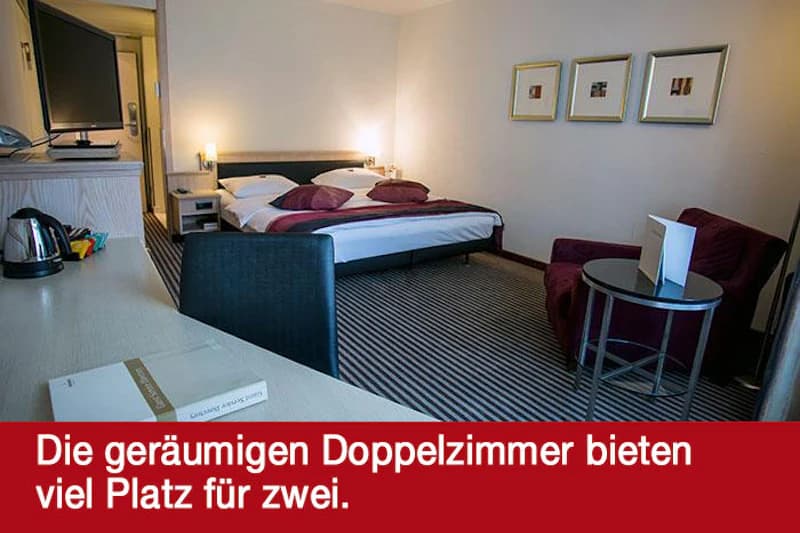 Hotelzimmer mit Doppelbett, Schreibtisch, Fernseher, Sessel und gestreiftem Teppich.