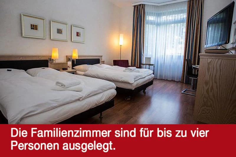 Hotelzimmer mit zwei Betten nebeneinander, Nachttischlampen, Fernseher, Schreibtisch und großem Fenster mit Gardinen.
