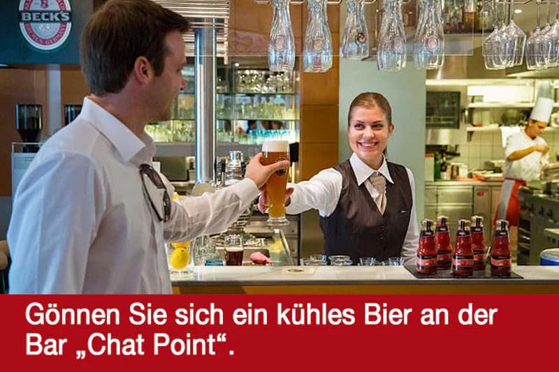 Servicekraft reicht einem Mann an der Hotelbar ein Glas Bier; Gläser hängen, Flaschen und Koch im Hintergrund.