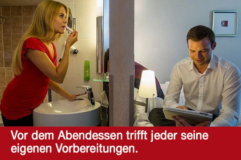 Frau putzt sich die Zähne am Waschbecken, nebenan sitzt ein Mann auf dem Bett mit Tablet, Zimmer durch Trennwand geteilt.
