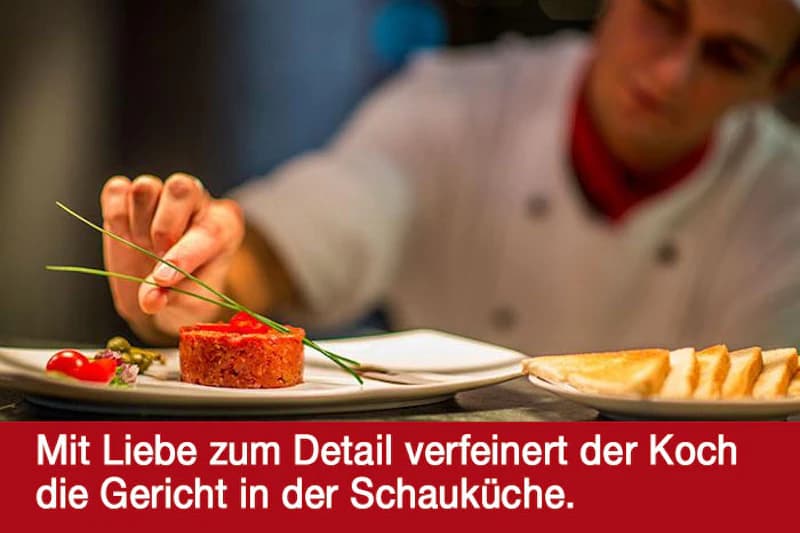Koch im Hintergrund richtet mit Hand ein Tatar auf einem Teller, garniert es mit Schnittlauch, daneben Toastscheiben.