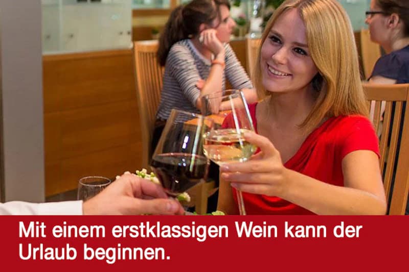Frau in rotem Kleid stößt mit Weißwein gegen ein Glas Rotwein in einem Restaurant mit Holzmöbeln an.