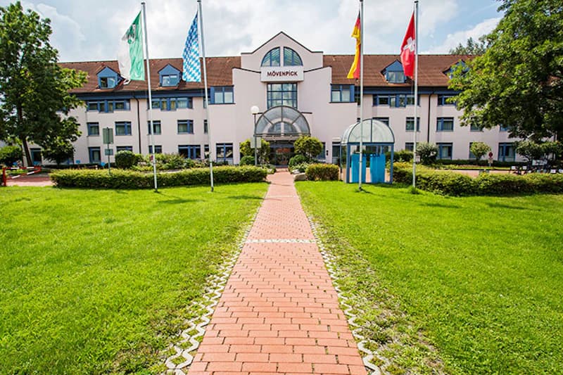 Zentraler roter Gehweg auf grüner Wiese zum dreistöckigen Mövenpick Hotel mit Glas-Eingang und mehreren Fahnen.