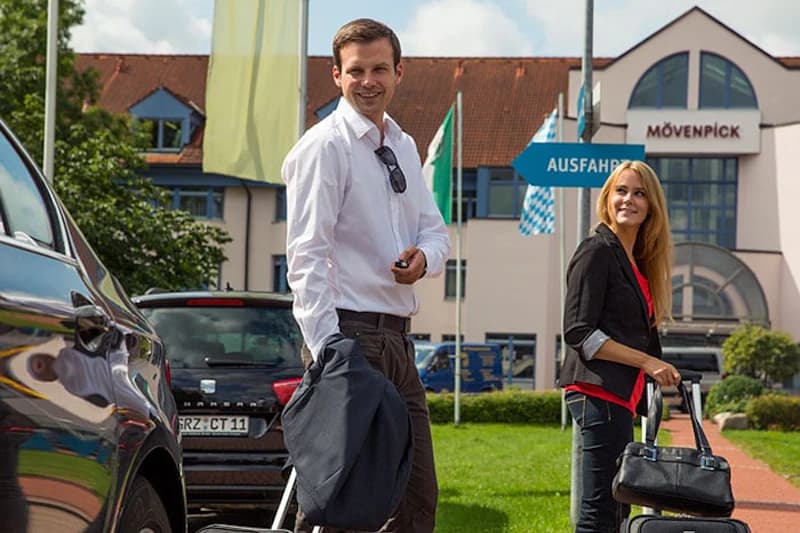 Mann und Frau mit Rollkoffern vor dem Mövenpick Hotel am Flughafen München, Auto im Vordergrund.