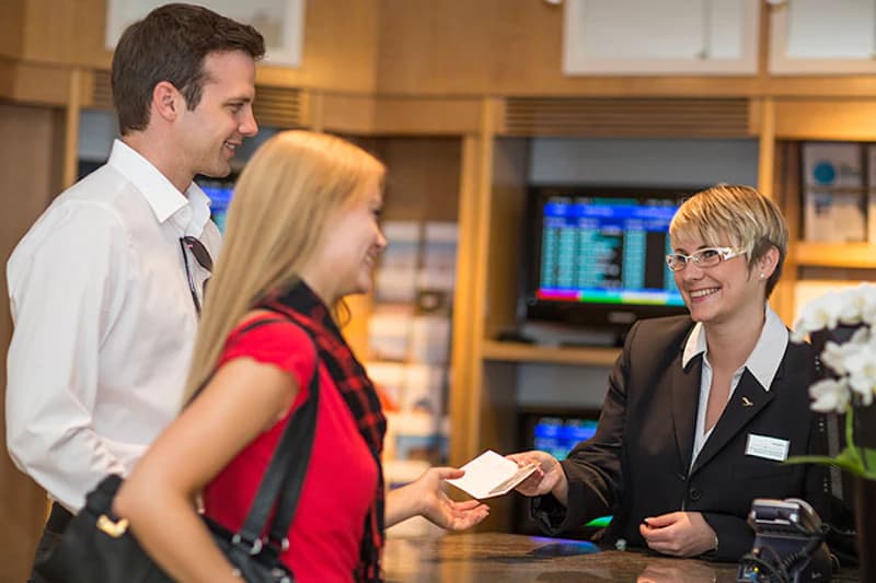 Hotelrezeption mit Empfangsmitarbeiterin, die zwei Gästen einen Zimmerschlüssel überreicht.
