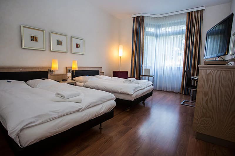 Hotelzimmer mit zwei Einzelbetten, Handtüchern auf den Betten, Nachttischlampen und großem Fenster mit Vorhängen.