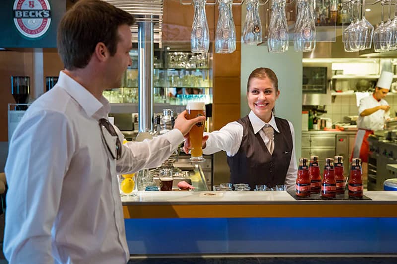 Frau hinter Bar reicht einem Mann ein Bierglas, Bar mit Hängengläsern und Koch im Hintergrund.